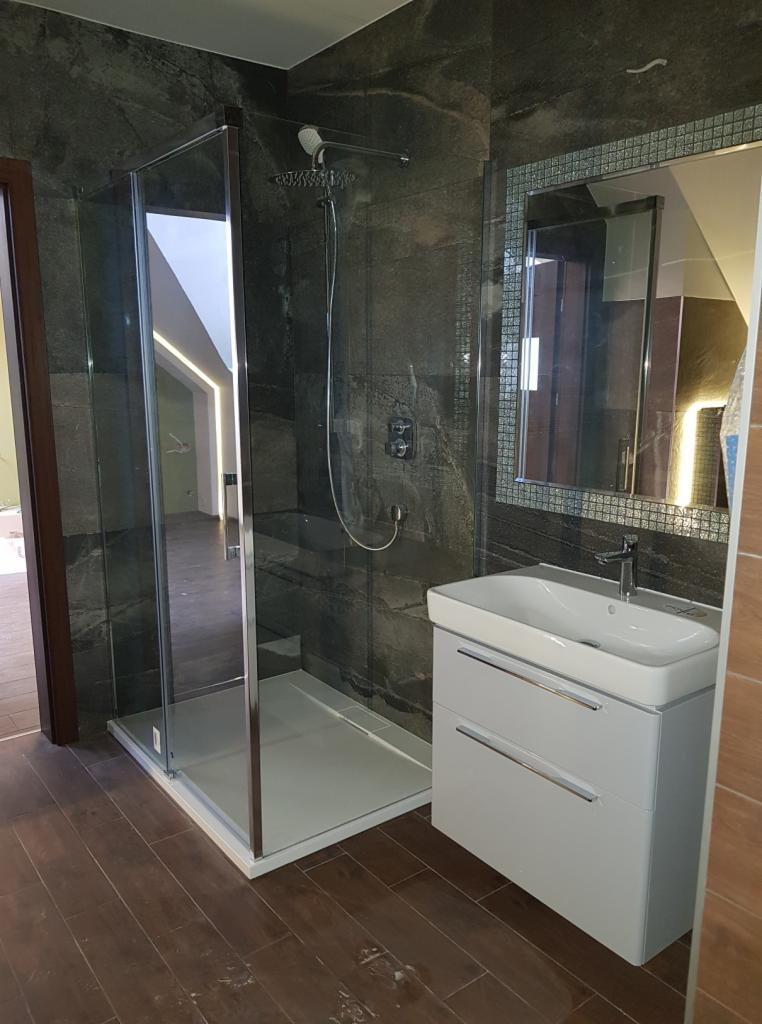 Nowoczesna łazienka z kabiną prysznicową typu walk-in, umywalką z szafką w kolorze białym i lustrem w mozaikowej ramie na tle ściany wyłożonej ciemnym kamieniem.