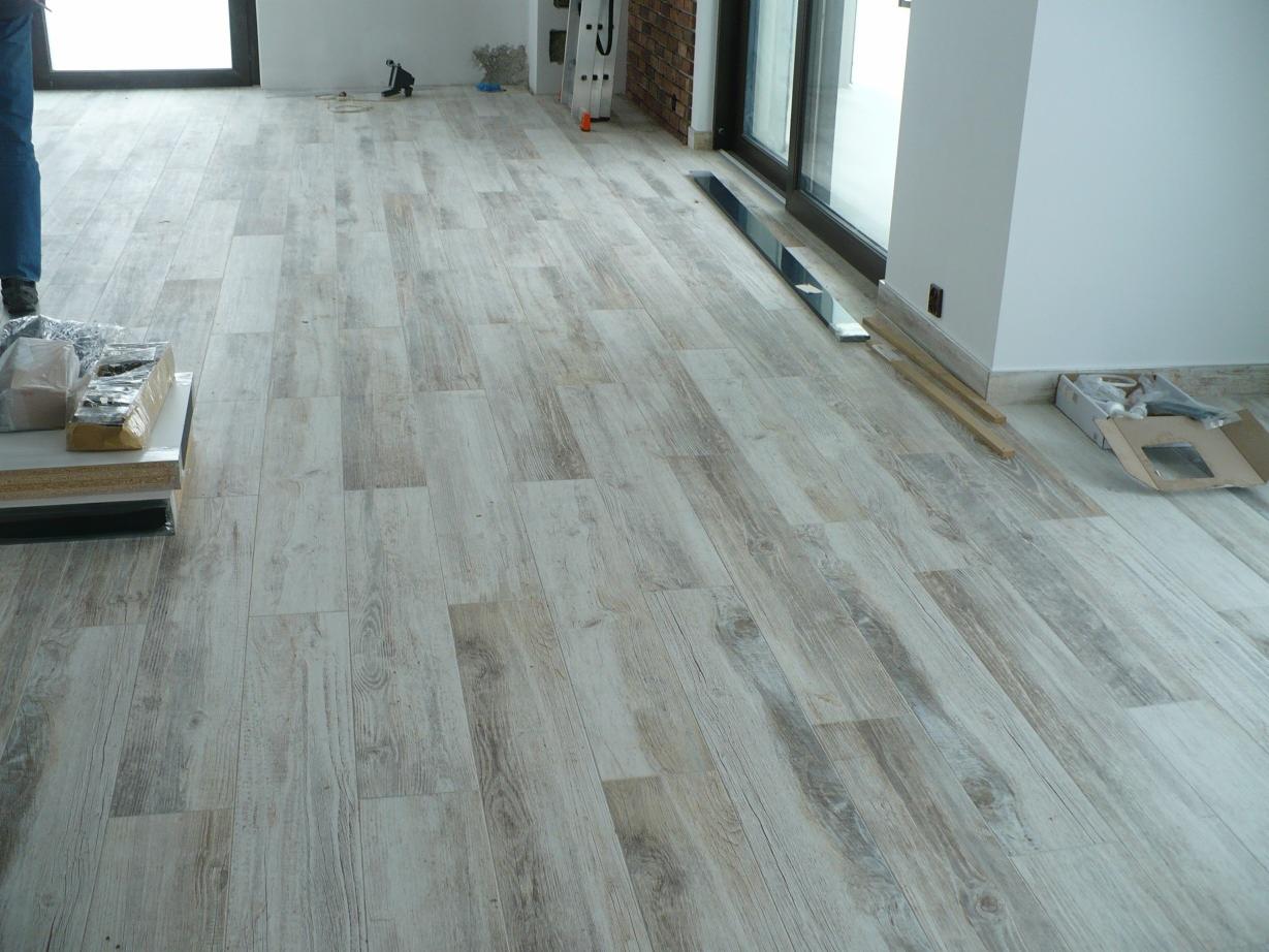 Nowo ułożona podłoga z paneli imitujących drewno w jasnych odcieniach, widoczne akcesoria montażowe i listwy przypodłogowe, fragment ściany z cegły i okno balkonowe.