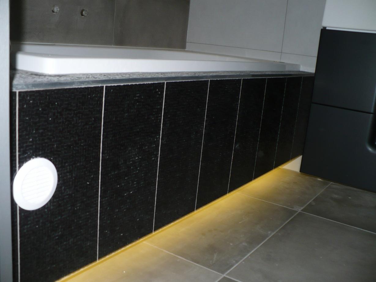 Obudowa wanny wykończona czarnymi, mozaikowymi płytkami z białymi fugami, podświetlona od dołu ciepłym, żółtym światłem LED, z widoczną kratką wentylacyjną i fragmentem szarej podłogi z płytek.