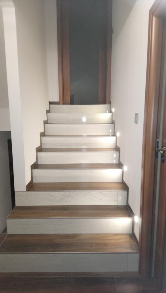 Schody wykończone panelami imitującymi drewno z subtelnym oświetleniem punktowym w ścianie, prowadzące do ciemnego pomieszczenia.