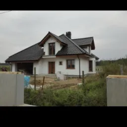 Nowo wybudowany dom z białym tynkiem, ciemną dachówką i drewnianą stolarką okienną, widoczny w tle fragment lasu i elementy ogrodzenia.