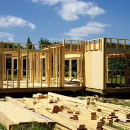 Konstrukcja szkieletowa domu w trakcie budowy, widoczne drewniane belki i słupy, przygotowane deski na pierwszym planie, błękitne niebo w tle.