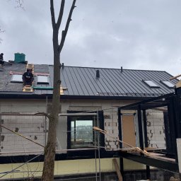 Superdekarze - Montaż czarnej blachy na dachu domu w trakcie budowy. Widoczni dekarze na rusztowaniu dachowym, okna dachowe i elementy konstrukcyjne budynku.