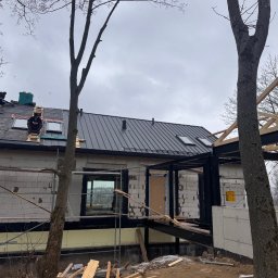 Superdekarze - Budowa domu w trakcie: montaż dachu z blachy na deskowaniu, widoczne okna dachowe i elementy więźby. Pracownicy na dachu. Surowy stan elewacji.