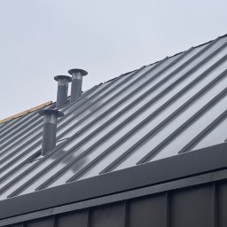 Superdekarze - Szary dach z blachy trapezowej z trzema kominami wentylacyjnymi na tle pochmurnego nieba. Widoczne krople deszczu na powierzchni dachu.