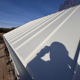 Superdekarze - Biała blacha trapezowa na dachu domu w świetle słonecznym, cień osoby robiącej zdjęcie. Widoczne rusztowanie i drzewa w tle.