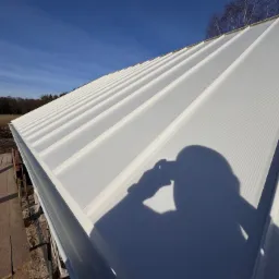 Biała blacha trapezowa na dachu domu w świetle słonecznym, cień osoby robiącej zdjęcie. Widoczne rusztowanie i drzewa w tle.