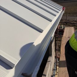 Superdekarze - Biały dach z blachy trapezowej z zamontowaną rynną, widok z góry z fragmentem pracownika w odblaskowej kamizelce na rusztowaniu. Słoneczny dzień.