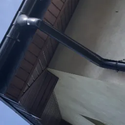 Widok z dołu na fragment elewacji budynku z zamontowanym, błyszczącym, czarnym systemem rynnowym z widocznymi mocowaniami. Widoczny fragment dachu z brązową podbitką.