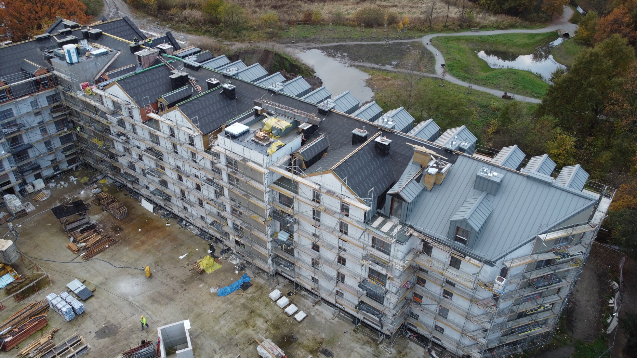 Wielopiętrowy budynek w trakcie budowy z częściowo ukończonym dachem z blachy w odcieniach szarości, otoczony rusztowaniami i materiałami budowlanymi.