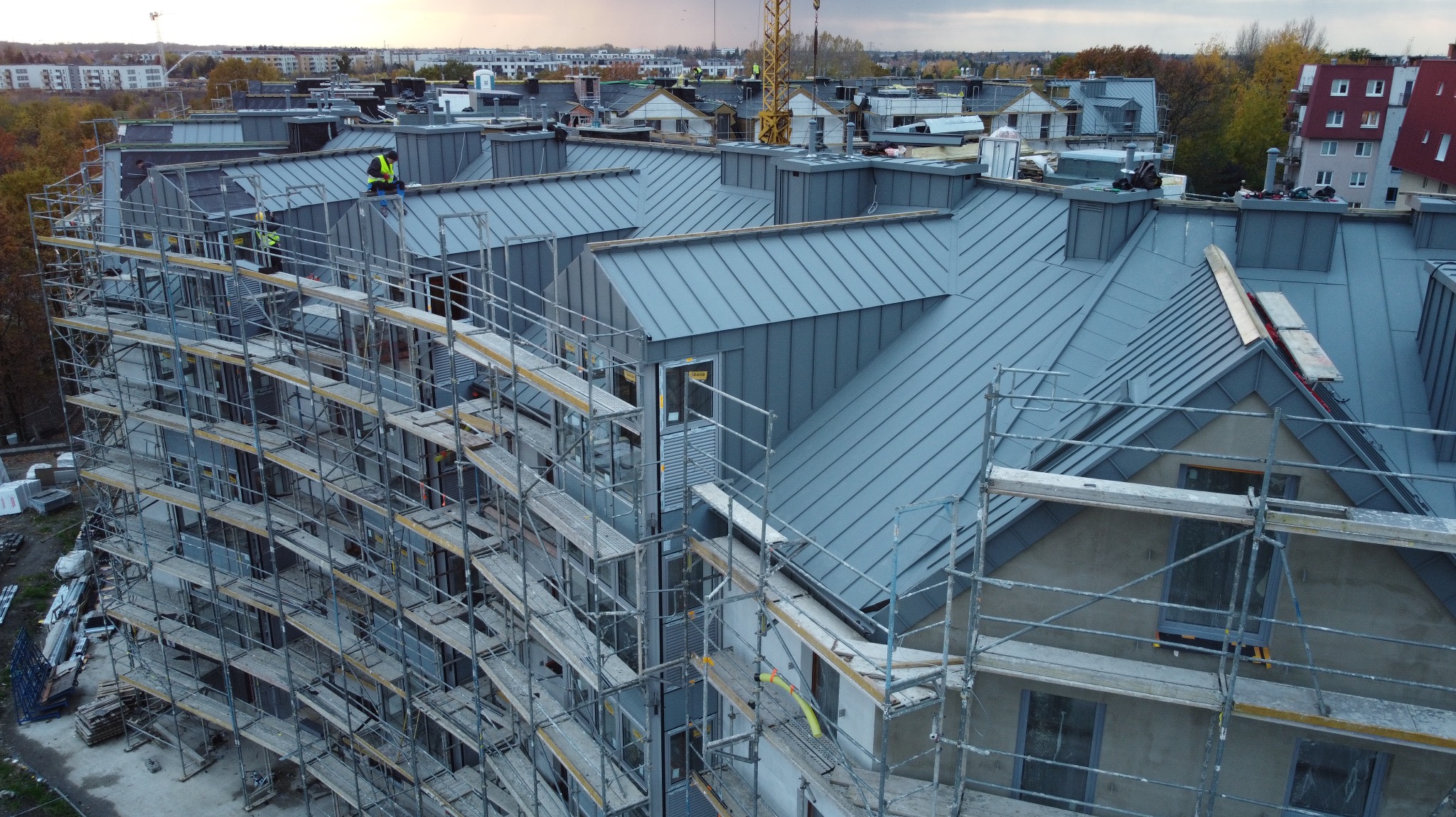Wielorodzinny budynek w trakcie krycia dachu blachą na rąbek stojący. Widoczne rusztowania, pracownicy w odblaskowych kamizelkach i dźwig w tle.
