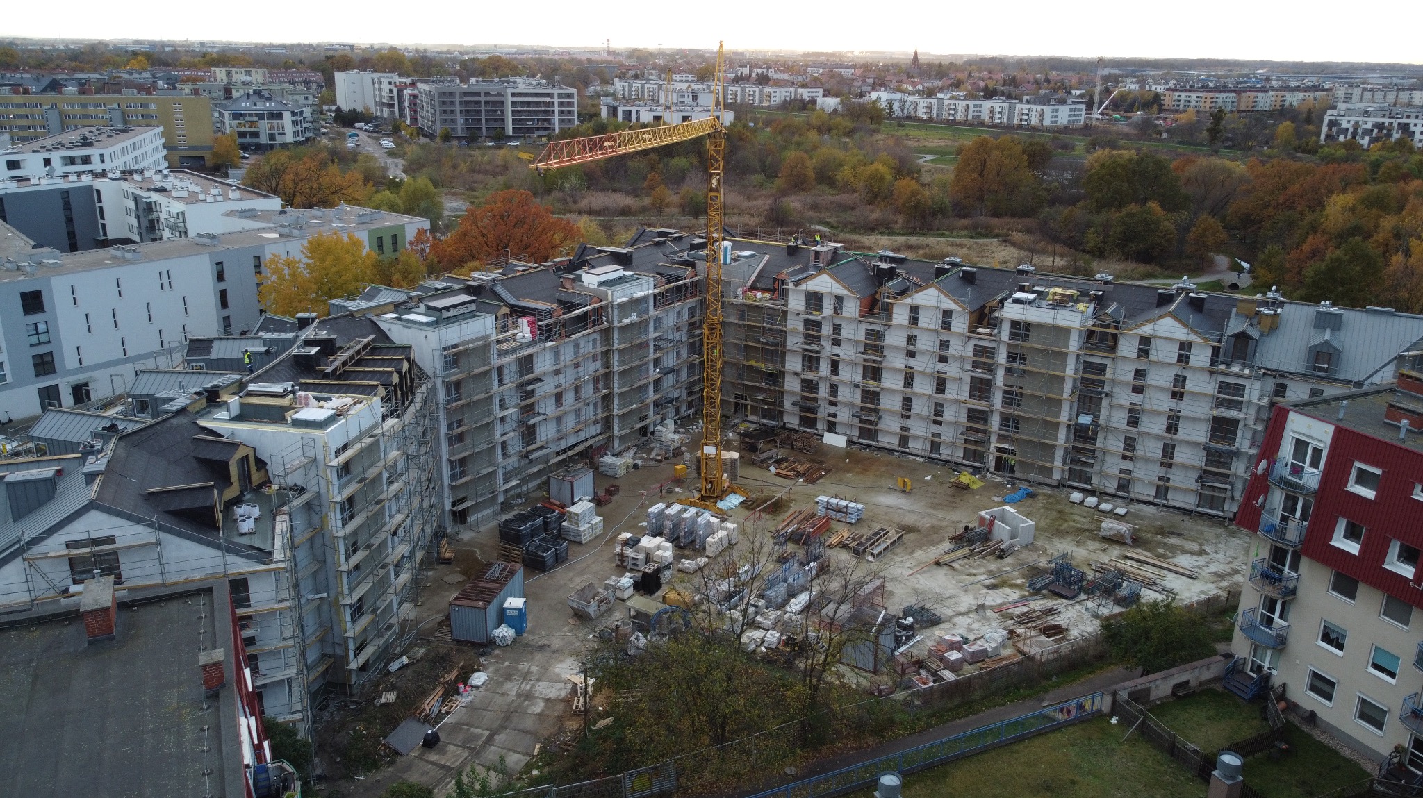 Budowa wielopiętrowego budynku z dachem w trakcie realizacji, widoczny dźwig budowlany i rusztowania. Prace dekarskie w toku, widok z góry na plac budowy.