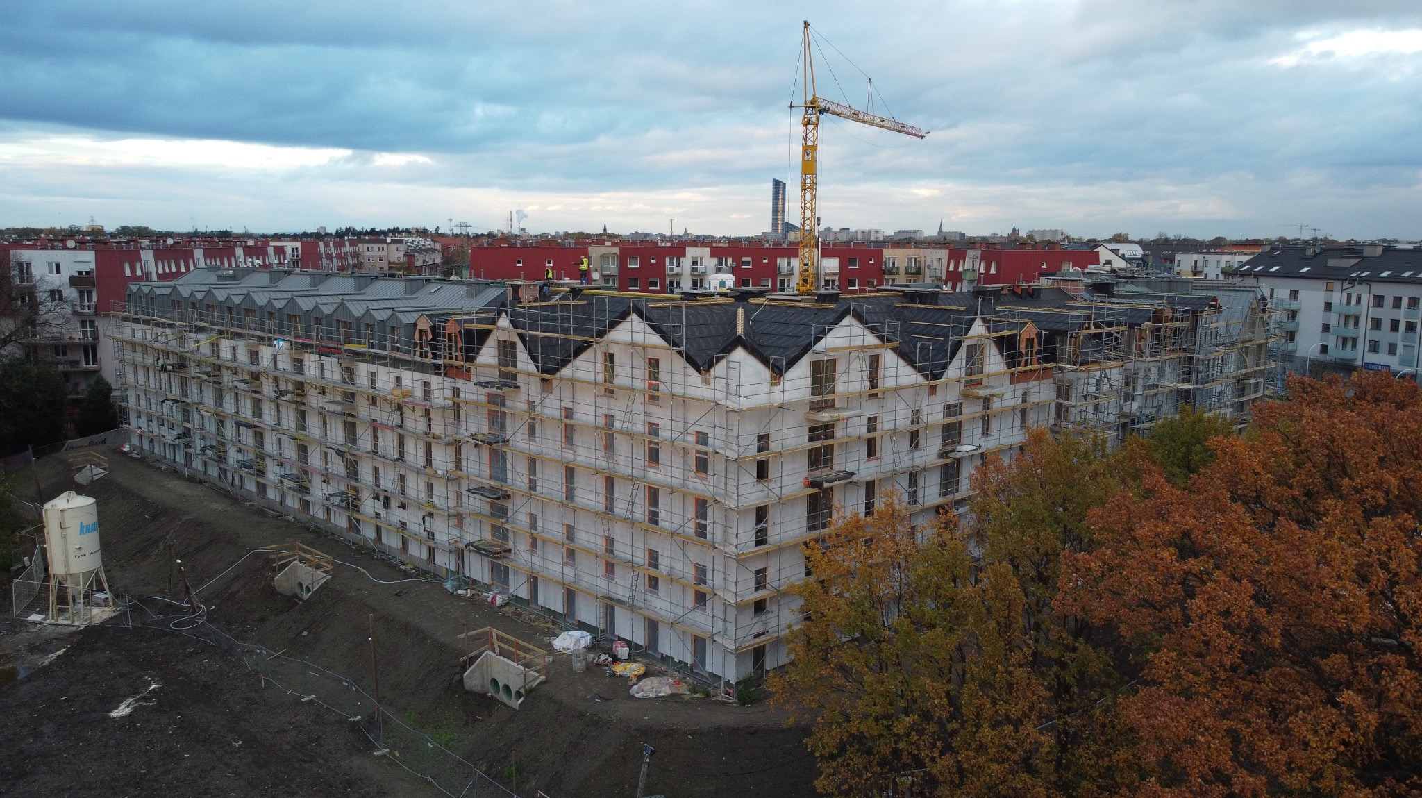 Wielopiętrowy budynek w trakcie prac dekarskich, z widocznymi rusztowaniami i dźwigiem w tle. Część dachu pokryta grafitową dachówką. Widok z góry, jesienne drzewa po prawej.
