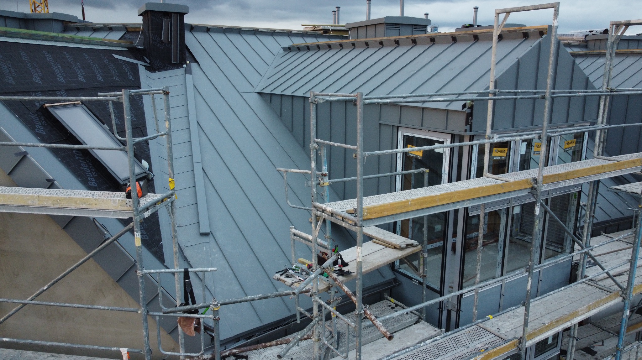 Szary dach z blachy na budynku mieszkalnym, widoczny z rusztowania. Fragment dachu pokryty papą. Okna z naklejkami DAKO. Pochmurne niebo.