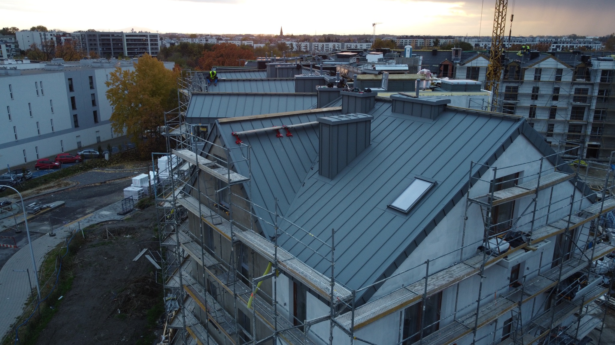 Nowoczesne krycie dachu blachą na budynku wielorodzinnym w trakcie prac. Widoczne rusztowania i pracownicy. W tle zabudowa miejska i zachodzące słońce.