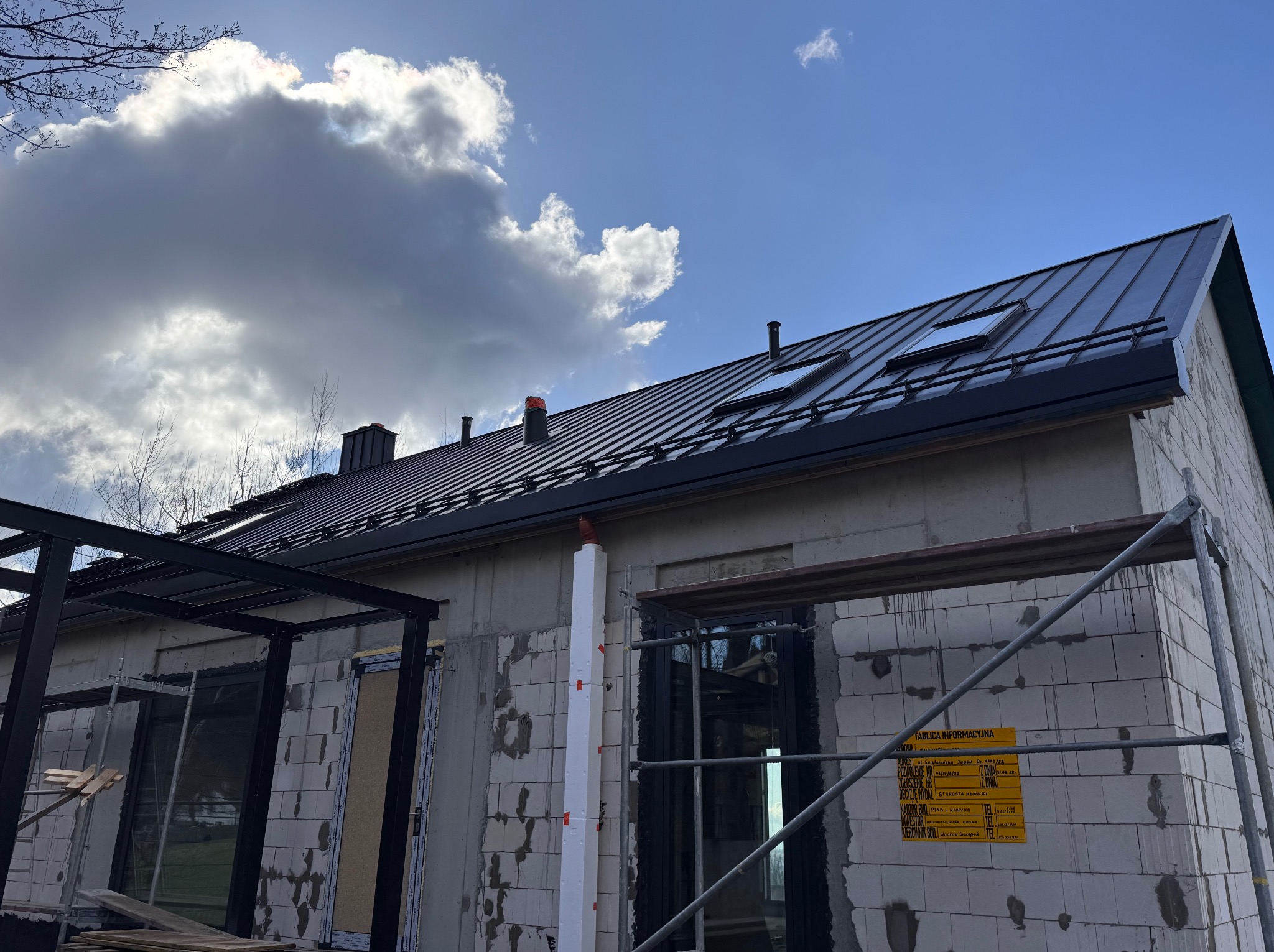 Dom w stanie surowym zamkniętym z czarnym dachem z blachy na tle błękitnego nieba z chmurami. Widoczne okna dachowe i elementy konstrukcyjne.