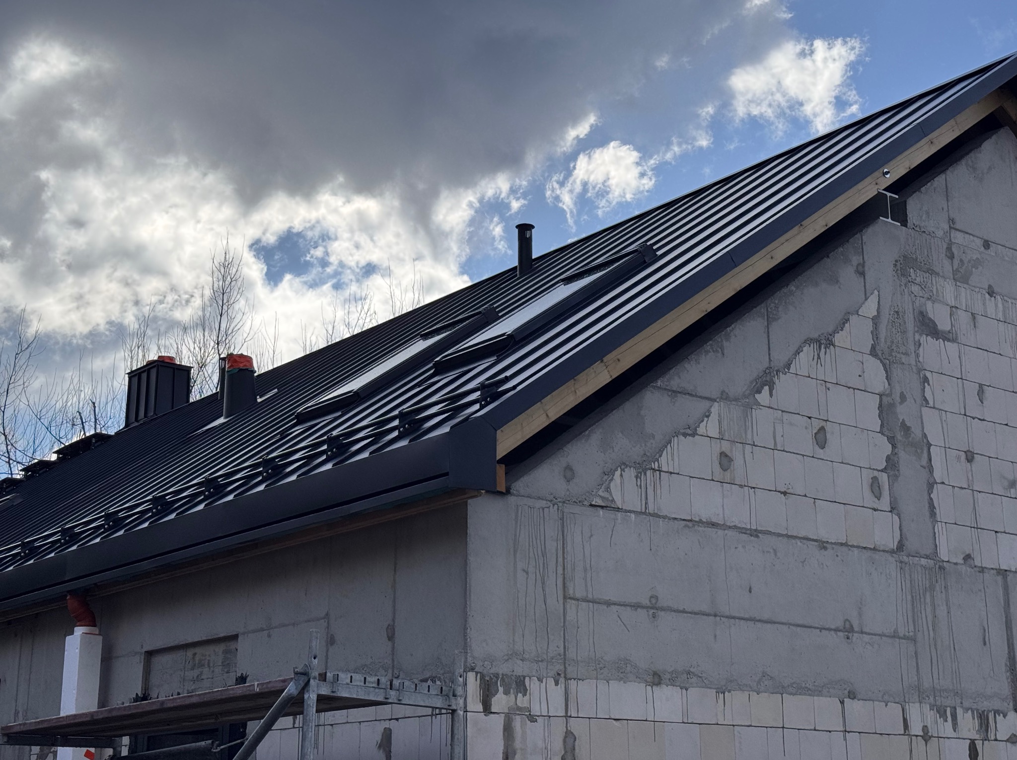 Czarny dach z blachy na surowym budynku z betonu komórkowego, widoczne okna dachowe i komin, pochmurne niebo w tle. Częściowo ukończona obróbka.