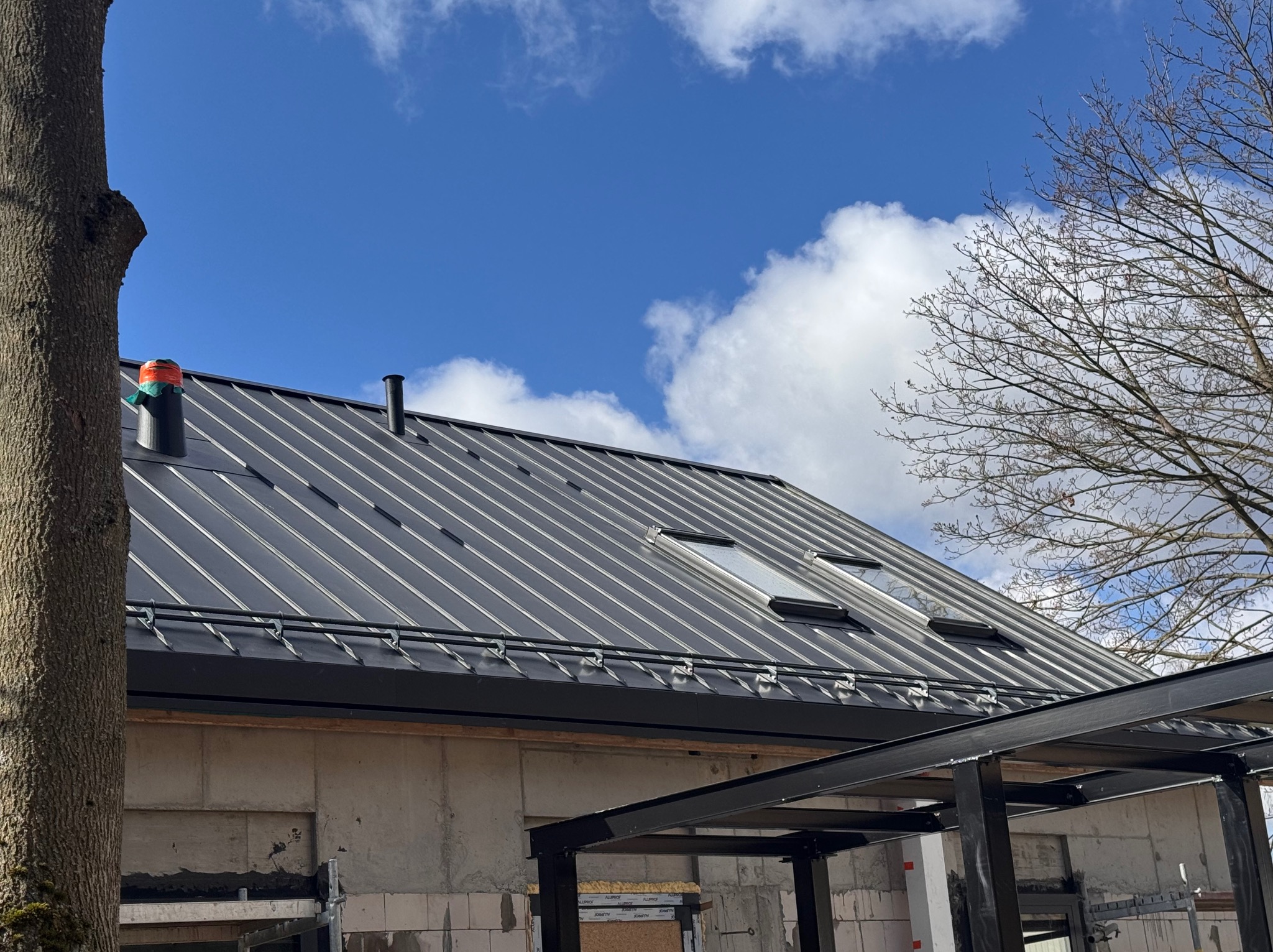 Szary dach z blachy na budynku w trakcie budowy, widoczne okna dachowe, komin z pomarańczowym zabezpieczeniem, błękitne niebo i fragment drzewa.