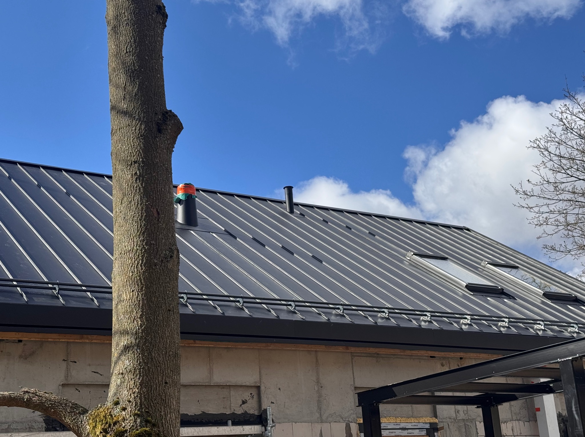 Nowoczesny dach z blachy na budynku w trakcie budowy, widoczne okna dachowe i komin, na pierwszym planie pień drzewa.