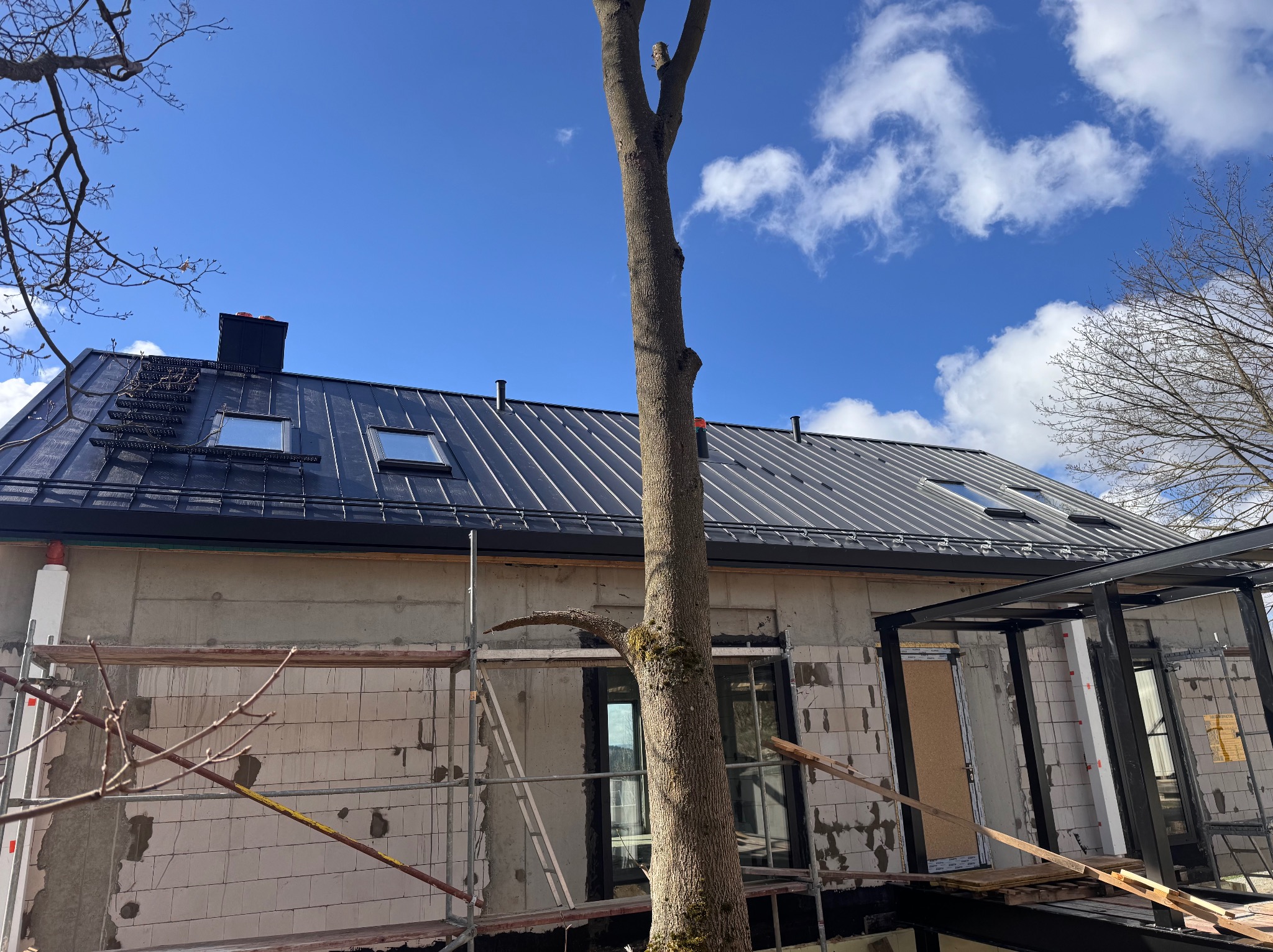 Dom w trakcie budowy z czarnym dachem z blachy trapezowej i oknami dachowymi, widoczne rusztowania i drzewo na pierwszym planie, błękitne niebo w tle.