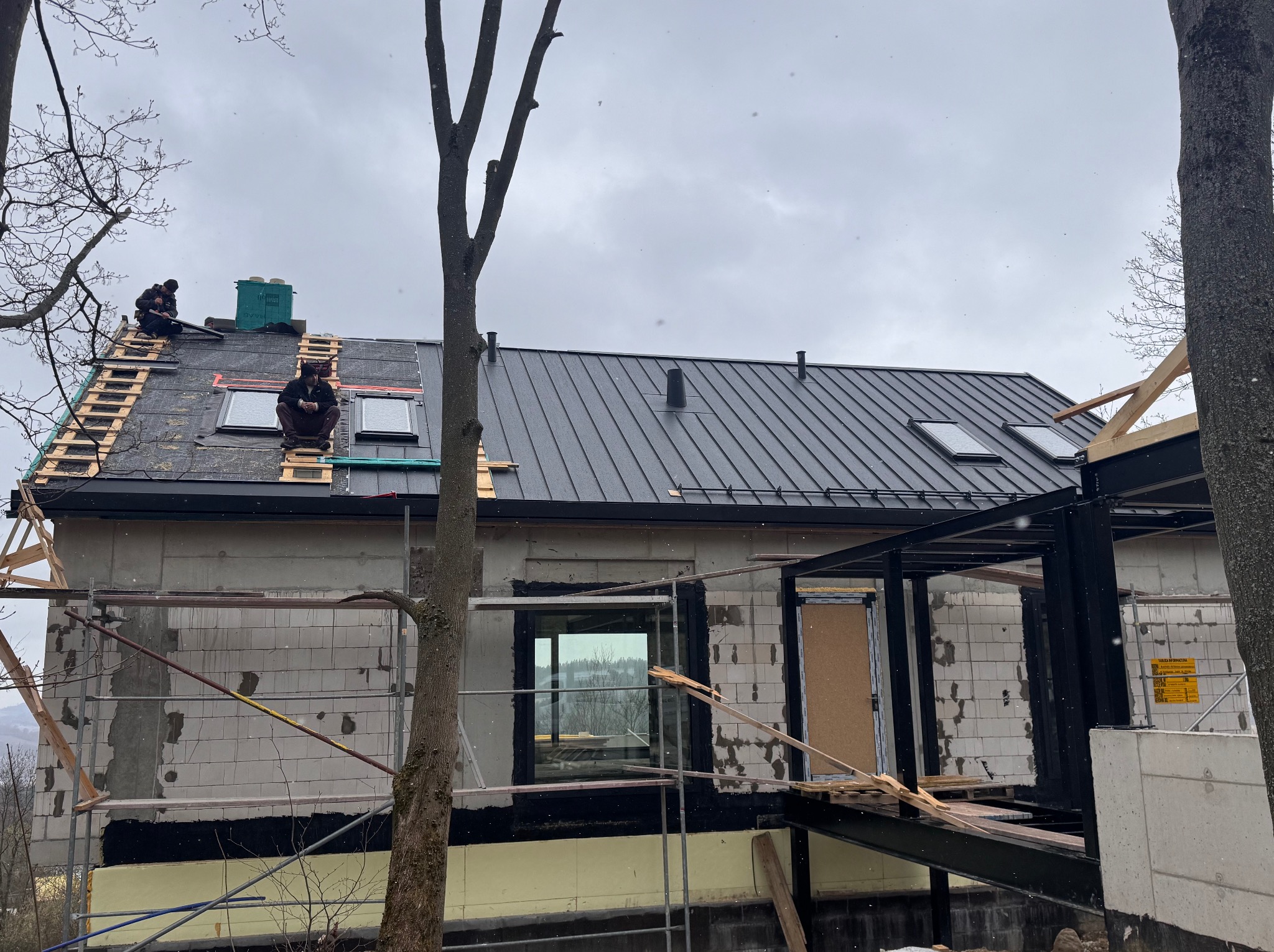 Montaż czarnej blachy na dachu domu w trakcie budowy. Widoczni dekarze na rusztowaniu dachowym, okna dachowe i elementy konstrukcyjne budynku.