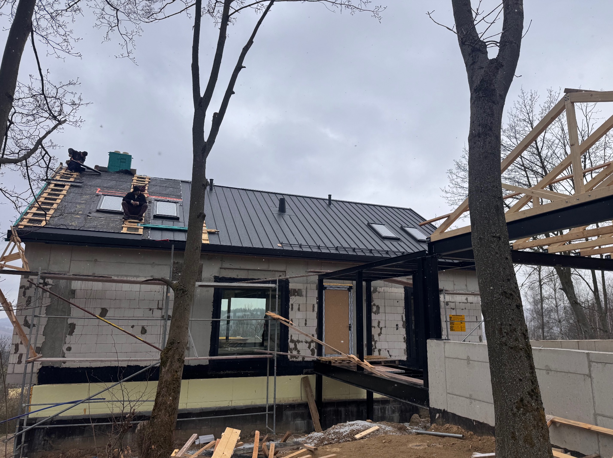 Budowa domu w trakcie: montaż dachu z blachy na deskowaniu, widoczne okna dachowe i elementy więźby. Pracownicy na dachu. Surowy stan elewacji.