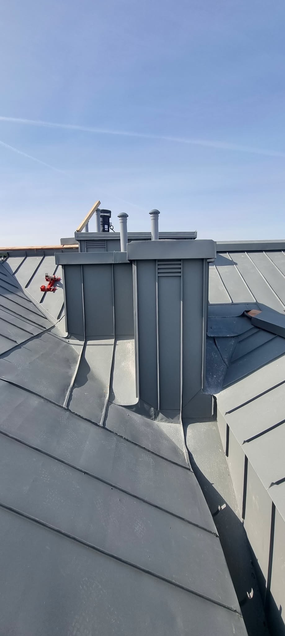 Szary dach z blachy na rąbek stojący z kominem i wentylacją, widok z góry na tle błękitnego nieba. Elementy montażowe widoczne na dachu.