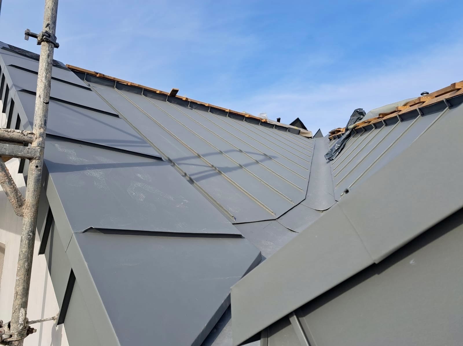 Szary dach z blachy na tle błękitnego nieba, widoczne elementy montażowe i rusztowanie. Ujęcie z dołu, podkreślające geometrię dachu.