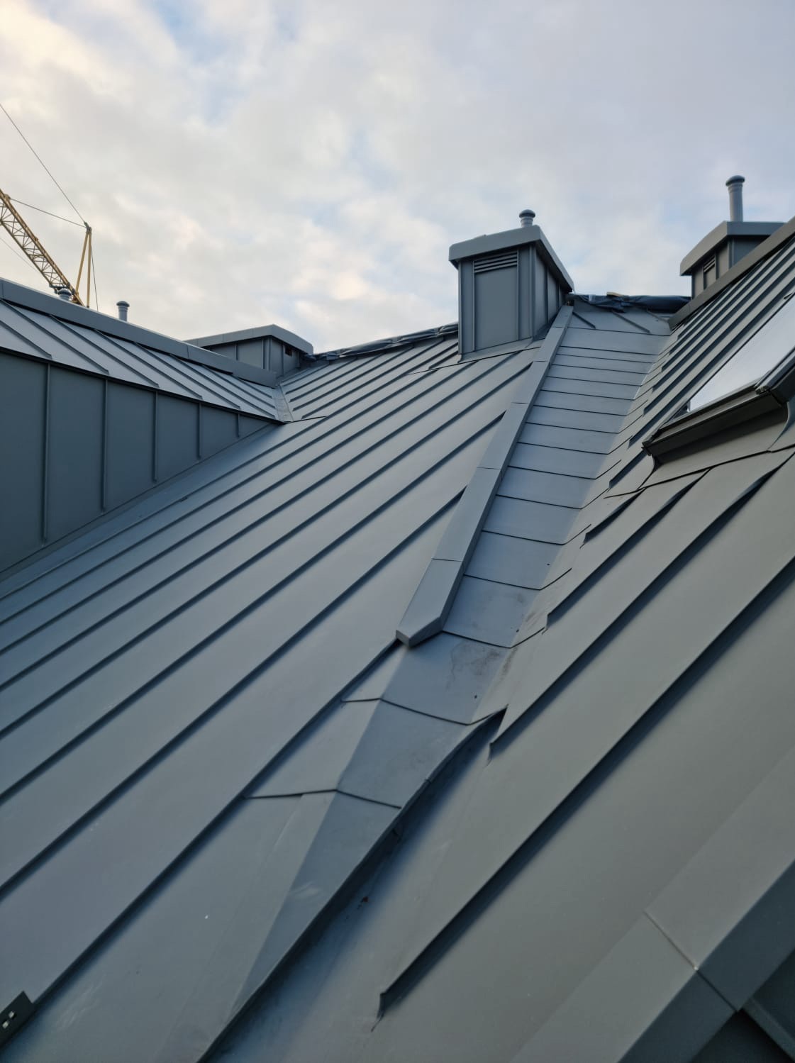 Szary dach z blachy na budynku, widoczne kominy i okno dachowe, w tle fragment żurawia budowlanego i pochmurne niebo.