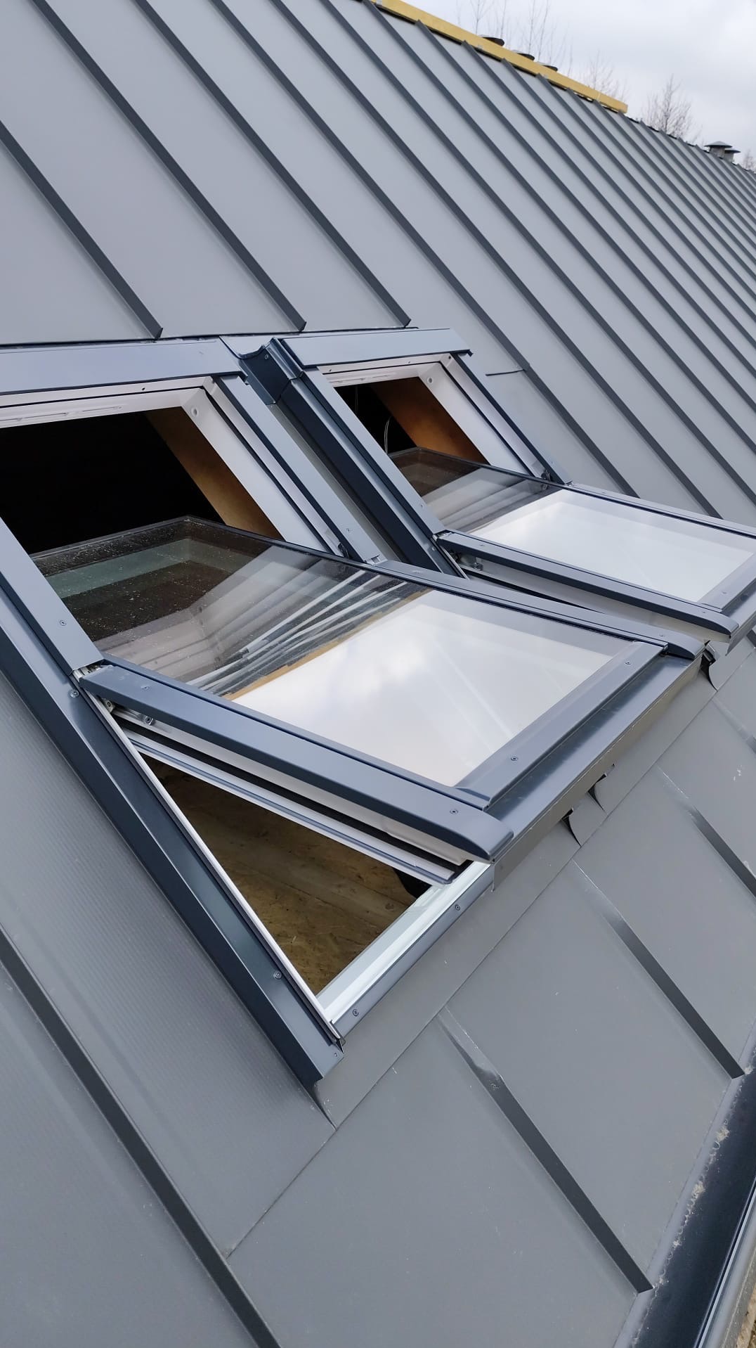 Szary dach z blachy na rąbek z dwoma oknami dachowymi, częściowo otwartymi. Widoczne drewniane elementy konstrukcji okien i blacha na dachu.