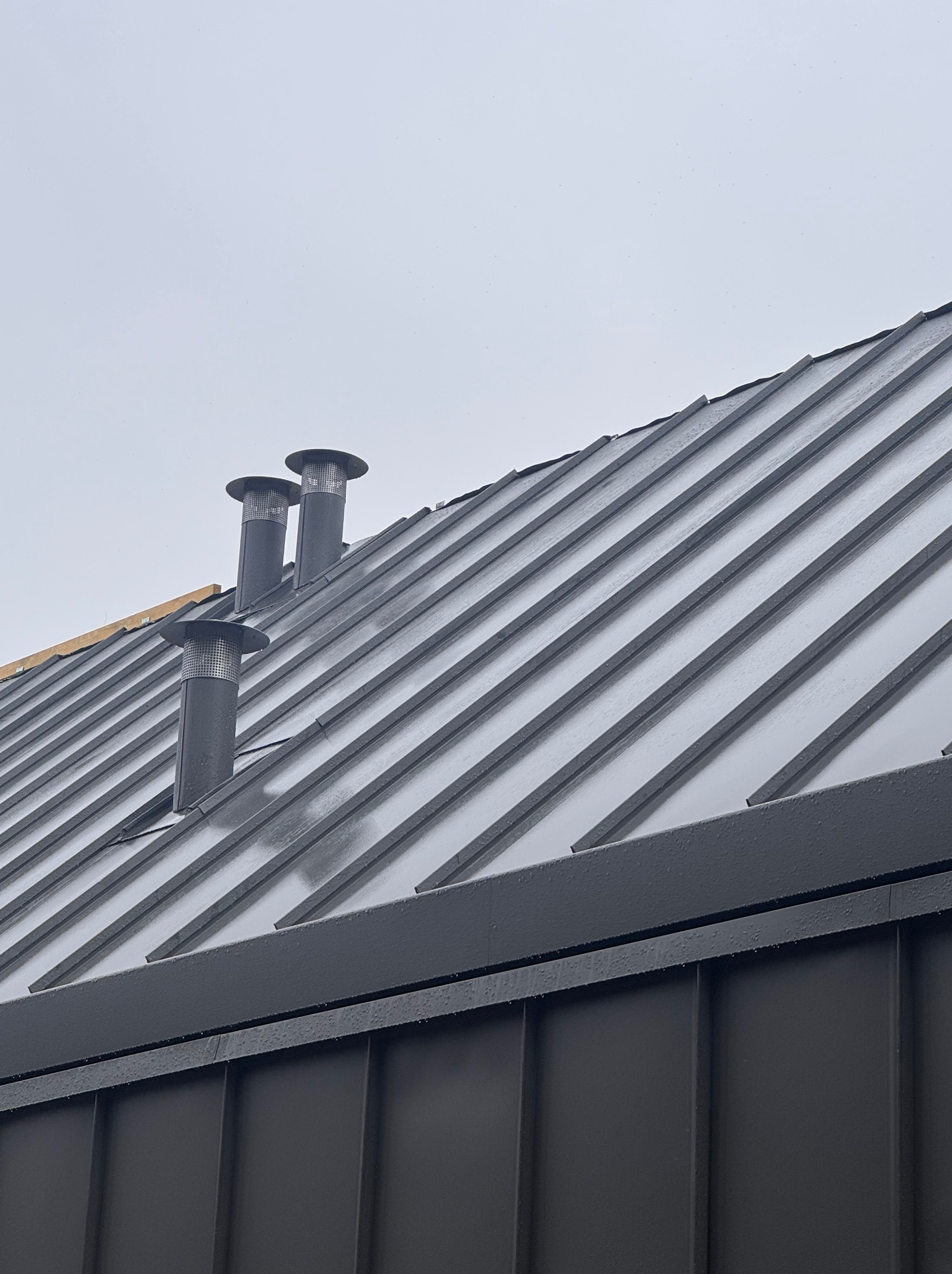 Szary dach z blachy trapezowej z trzema kominami wentylacyjnymi na tle pochmurnego nieba. Widoczne krople deszczu na powierzchni dachu.