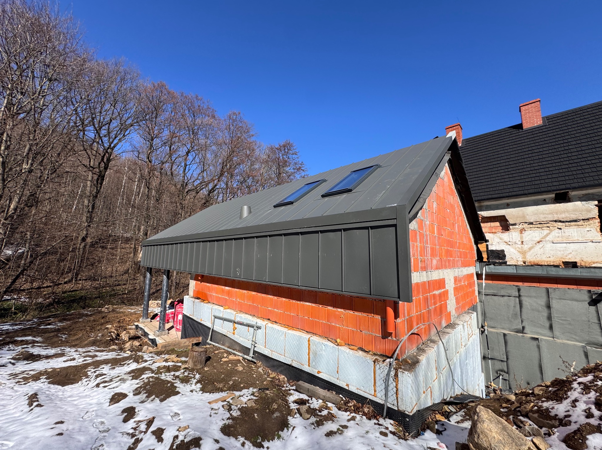 Budynek w trakcie budowy z szarą blachą na dachu i oknami dachowymi, widoczne czerwone cegły i ośnieżone podłoże.