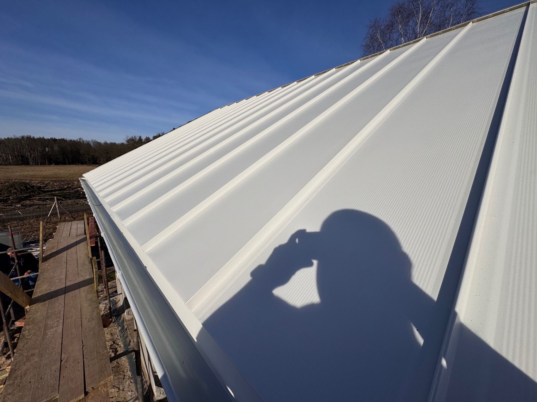 Biała blacha trapezowa na dachu domu w świetle słonecznym, cień osoby robiącej zdjęcie. Widoczne rusztowanie i drzewa w tle.