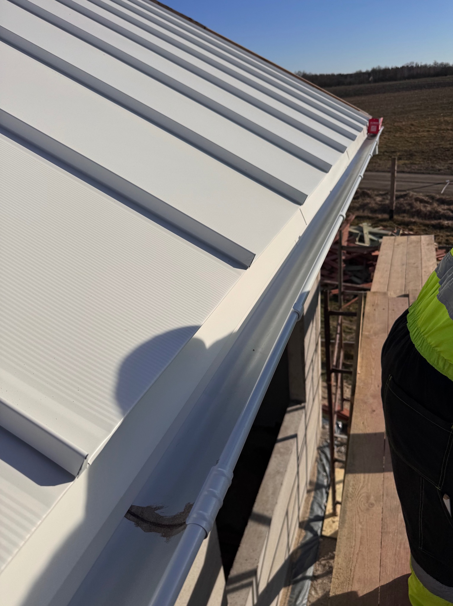 Biały dach z blachy trapezowej z zamontowaną rynną, widok z góry z fragmentem pracownika w odblaskowej kamizelce na rusztowaniu. Słoneczny dzień.