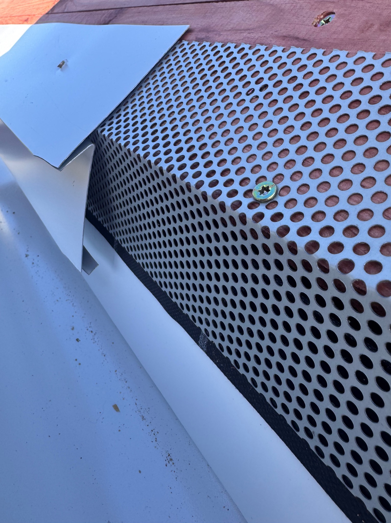 Szczegółowe ujęcie fragmentu dachu z metalową siatką wentylacyjną i elementami obróbki blacharskiej, mocowanymi śrubami, na tle izolacji.