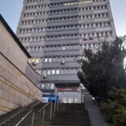 Wysoki, szary budynek biurowy widziany z dołu schodów wejściowych, z zaparkowanym radiowozem policyjnym przed wejściem.