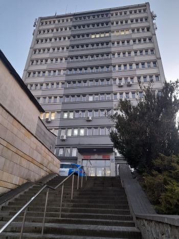 Wysoki, szary budynek biurowy widziany z dołu schodów wejściowych, z zaparkowanym radiowozem policyjnym przed wejściem.