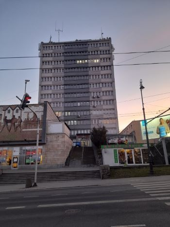 Wielopiętrowy budynek biurowy z antenami na dachu, sfotografowany z poziomu ulicy o zmierzchu, z widocznymi schodami prowadzącymi do wejścia i muralem po lewej stronie.