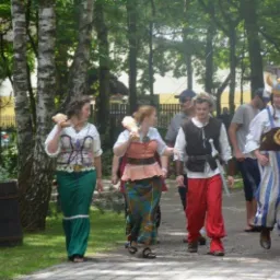 Grupa osób w kostiumach stylizowanych na historyczne, idąca ścieżką w parku, z rekwizytami imitującymi broń, w tle dmuchaniec i drzewa.