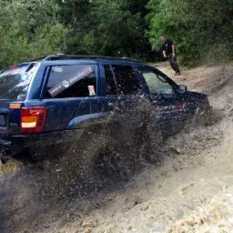 Niebieski samochód terenowy pokonuje błotnisty teren, w tle zalesiony obszar i osoba obserwująca.