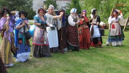 Grupa kobiet w barwnych, stylizowanych strojach ludowych, prawdopodobnie uczestniczących w imprezie tematycznej na trawniku przed budynkiem, jedna z nich trzyma mikrofon.