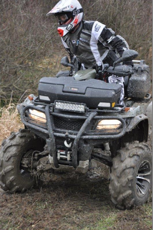 Mężczyzna w kasku i goglach na quadzie pokonuje błotnisty teren, widoczne oświetlenie LED na pojeździe.