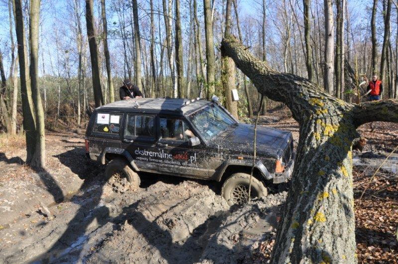 Czarne auto terenowe z napisem 'ekstremalne.pl' zakopane w błocie na leśnym terenie podczas imprezy off-road, z osobą stojącą na dachu i pochylonym drzewem w kadrze.