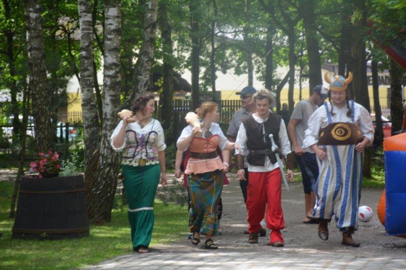 Grupa osób w kostiumach stylizowanych na historyczne, idąca ścieżką w parku, z rekwizytami imitującymi broń, w tle dmuchaniec i drzewa.