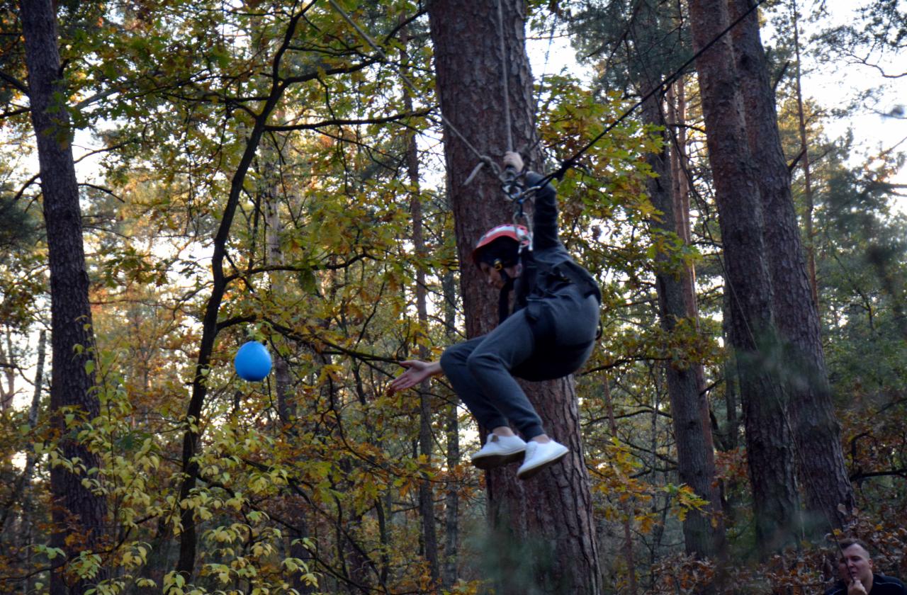Osoba w kasku i sportowym stroju zjeżdża na linie w parku linowym, z boku widoczny niebieski balon.