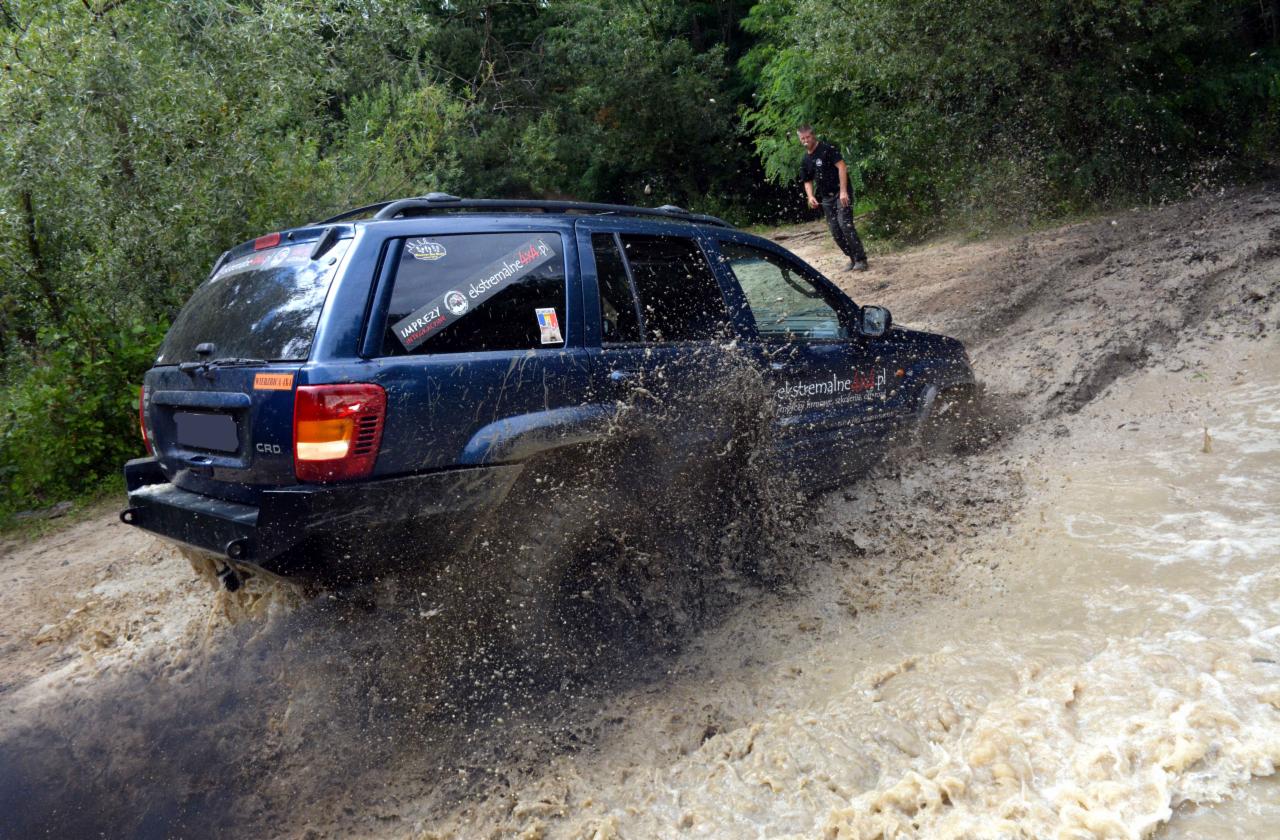 Niebieski samochód terenowy pokonuje błotnisty teren, w tle zalesiony obszar i osoba obserwująca.