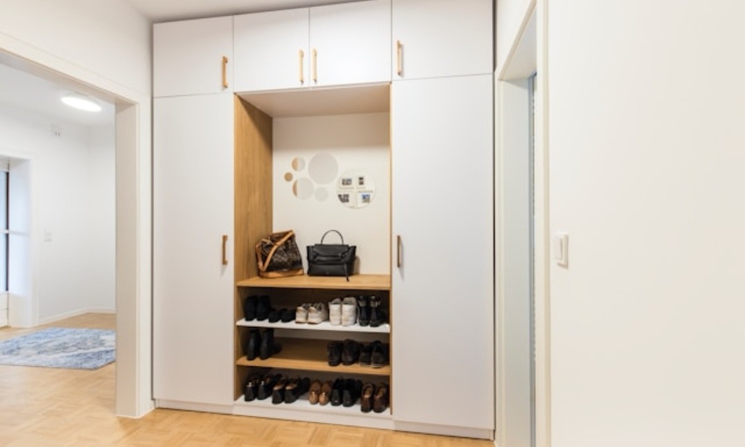 Biała szafa wnękowa z drewnianymi akcentami i półkami na buty, uzupełniona damskimi torebkami, na podłodze parkiet w jodełkę.