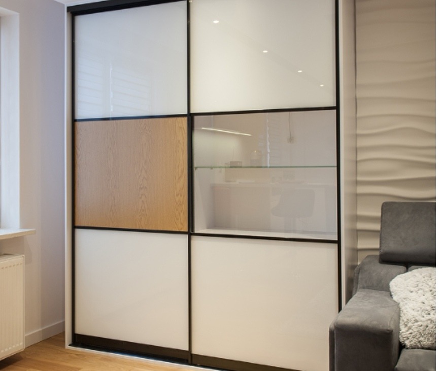 Nowoczesna szafa przesuwna z białymi panelami, jednym panelem z okleiny drewnopodobnej i przeszkloną witryną, czarne ramy, widoczny fragment sofy i dekoracyjna ściana w tle.