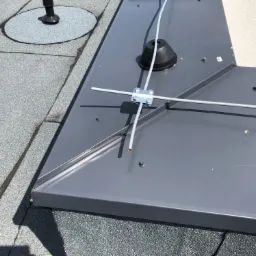 Szary, płaski dach pokryty papą z metalową obróbką komina i mocowaniem odgromowym, widok z góry.