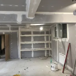 Wnętrze w trakcie prac wykończeniowych: ściany z płyt kartonowo-gipsowych, szpachlowane łączenia, drewniane p&oacute;łki wnękowe, widoczne narzędzia i wiadro z farbą.